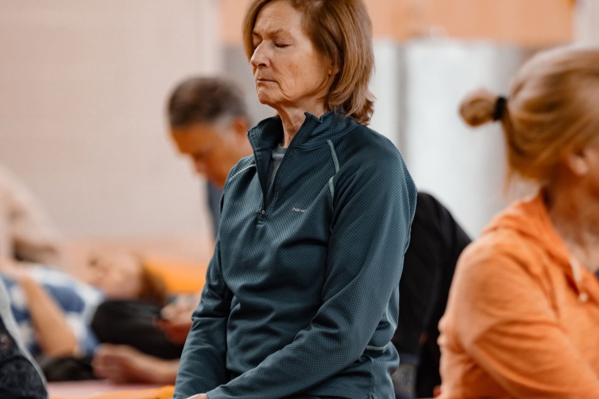 A room of students seated upright on orange bolsters in a Boulder practice space, eyes closed, mid-practice